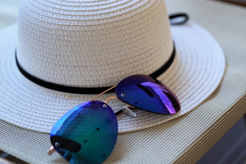 A white sunhat with a black ribbon on a table with a pair of blue lensed sunglasses